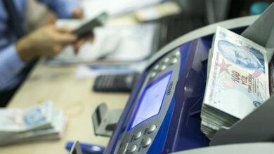 Para sayma makinesinin üzerinde 100 TL'lik banknotlar var. Fotoğrafta, flu bir şekilde hesap makinesi, 100 TL'lik banknotlar, bilgisayar ekranı ve klavyesi, masanın üzerinde duruyor. Aynı zamanda yüzü gözükmeyen bir kişi elindeki paraları sayıyor.