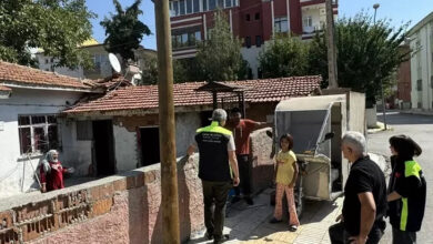 Tek katlı bir evin önünde 3 tekerli motosiklet var. 3 tekerli motosikletin önünde genç bir erkek, yeşil renkli bir üniforma giymiş başka bir erkek ile konuşuyor. Arka taraflarında üniforma giymiş biri kadın, üniformasız bir erkek daha var. Aynı zamanda evin bahçesinde yaşlı bir kadın daha var.