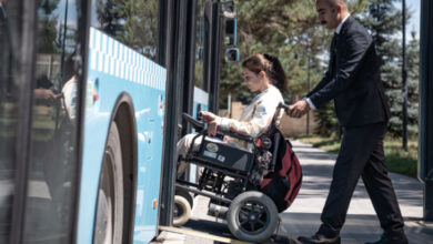 Akülü tekerlekli sandalye kullanan engelli bireyi, belediye otobüsü şoförü engelli rampasını açarak ve engelli bireyin binmesine yardımcı oluyor.