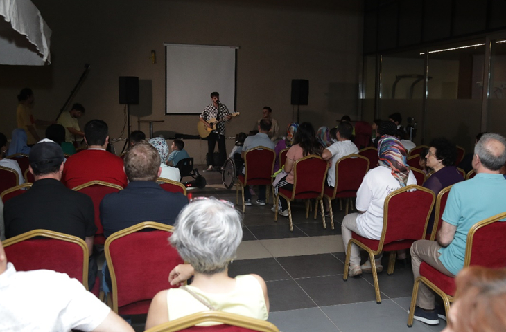 Fotoğrafta, elinde gitar tutup çalan bir müzisyen ve karşısında yer alan SMA’lı bireyler ve aileleri var.