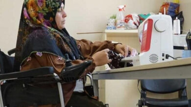 Akülü tekerlekli sandalye kullanıcısı Ayşe Pehlivan, bir masanın üzerinde bulunan dikiş makinesi ile elindeki kumaş parçasını dikiyor.