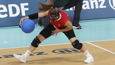 Golbol Kadın Milli Takımı sporcusu, gözünde siyah bir bant takılı ve elinde mavi renkli yuvarlak bir top var. Paralimpik sporcu yere doğru eğilerek topu atmaya hazırlanıyor.