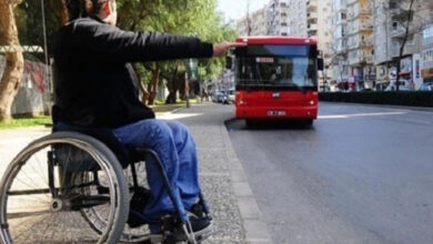 Tekerlekli sandalye kullanıcısı engelli birey, otobüs durağına doğru yaklaşan otobüse elini kaldırarak durması için işaret veriyor.