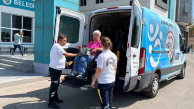 Akülü tekerlekli sandalye kullanıcısı engelli birey, bir minibüsün arkasına özel olarak yapılmış engelli asansörü ile araçtan aküllü sandalye üzerinde indiriliyor. Engelli bireyin yanında refakat ile belediye personelleri var.