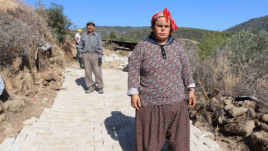 Engelli birey Emine Çolak ve ailesi taş yolun üstünde ayakta bir şekilde duruyor. Arka tarafta ise taş yolun döşemesine devam eden işçiler var.
