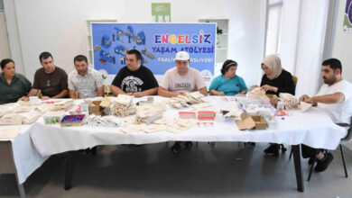 Engelli bireyler beyaz masa örtüsünün serili olduğu, yan yana duran birkaç masaların etrafında krem renginde bez parçalardan kalem kutusu yapıyorlar.