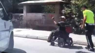 Fotoğrafta, bir polis ekip otosu ve akülü tekerlekli sandalye kullanan engelli bir bireyin arkasında yürüyen polis memuru var.