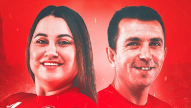 Para okçuluk branşında Sadık Savaş ve Merve Nur Eroğlu'ndan oluşan Türkiye Klasik Yay Karışık Milli Takımı sporcuları, kırmızı arka planı olan fotoğrafta hafif sırtları birbirine dönük bir şekilde duruyorlar ve gülümsüyorlar.