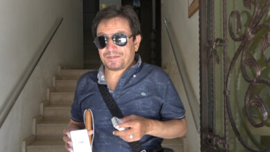 Siyah bir gözlük takan görme engelli birey, elinde beyaz baston, zil ve uzaktan kumandayı tutuyor.