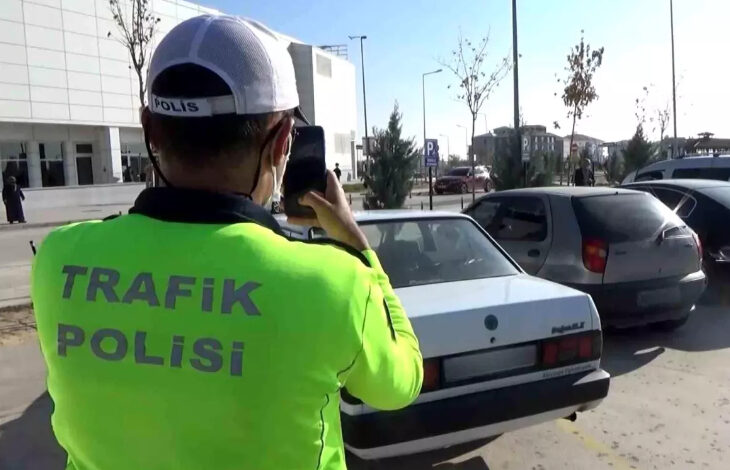 Fotoğrafta, bir otopark alanında trafik polisi sırtında yazan yeşil bir üniforma giymiş erkek otomobillerin fotoğrafını çekerek, hatalı park yapan araçlara ceza yazıyor.