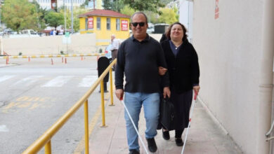 Ellerinde beyaz baston olan görme engelli kadın ve erkek kol kola girmiş, siyah gözlükleri takılı şekilde yürüyorlar.