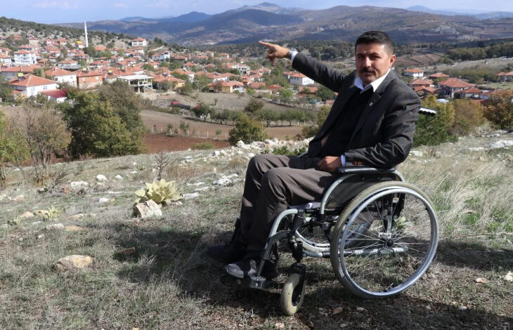 Tekerlekli sandalye üzerinde oturan Alan Köyü muhtarı Halil Yerlikaya, hizmet verdiği köyü görebileceği bir alandan bir elinin parmağını kaldırarak köyün olduğu yeri gösteriyor.