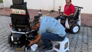 Fotoğrafta, üzerinde kimsenin oturmadığı akülü tekerlekli sandalyenin tekerlerinin olduğu bölüm ve ikisinin bağlantı noktasını bir kişi eğilerek kontrol ediyor. Aynı zamanda başka bir akülü tekerlekli sandalye üzerinde tamir için sıra bekleyen engelli birey duruyor.