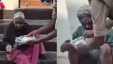 Fotoğrafta yer alan yaşlı çift bir taş üzerinde oturuyor ve görevli kişiler görme engelli çifte yemek yedirmeye çalışıyor.