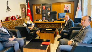 Fotoğrafta, bir makam odasında yer alan 4 kişi var. Makam koltuğu boş, makamın önündeki koltuklarda bir kadın, üç erkek oturuyor.