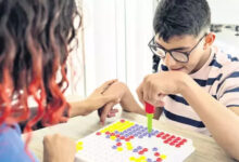 Engelli bir birey, önündeki kutuya renkli boncukları yerleştiriyor. Karşısında duran özel eğitim öğretmeni ise engelli öğrenciye destek oluyor.