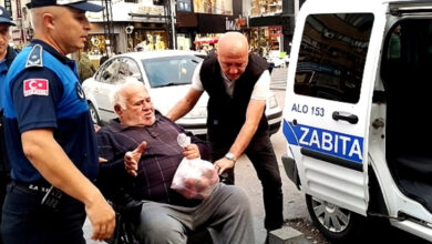Fotoğrafta, akülü tekerlekli sandalye üzerinde oturan yaşlı bir bireyin zabıta aracına doğru sandalyesinin sürüldüğü görülmektedir.