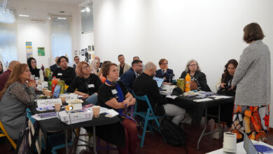 Yan yana katılmış masaların etrafındaki sandalyelerde kalabalık bir insan grubu oturuyor. Karşılıklı olacak şekilde iki grup şeklinde oturan kişiler karşılarındaki eğitmeni dinliyorlar.