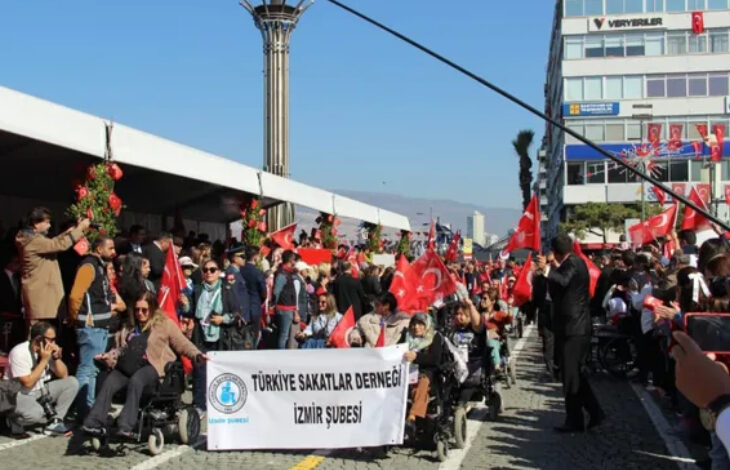 Tekerlekli sandalye kullanan engelli bireyler, bir dar sokaktan kortej ile birlikte ilerlerken en önde yer alan engelli bireyler “Türkiye Sakatlar Derneği İzmir Şubesi” yazılı bir pankart açıyor. Ellerinde Türk Bayrağı olan engelli bireyler sokak boyunca geçiş yapıyor.