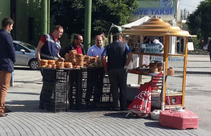 Fotoğrafta, simit camekanında dizili simit ve su şişeleri var. Simit satan kişi ve zabıta memurları da fotoğrafta yer alıyor.