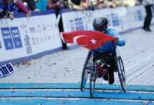 Fotoğrafta, önde bir ve yanlarda iki tekeri olan özel yapan tekerlekli sandalye ile engelli birey maraton koşusunu bitirirken Türk bayrağını çıkartarak bitiş çizgisini geçiyor.