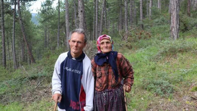Fotoğrafta, ağaçların ve küçük yeşil bitkilerin olduğu bir ormanlık alanda down sendromlu oğlu ile yan yana ellerindeki tahta bastona tutunarak ayakta duran anne Ayşe Aydoğan var.