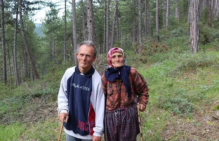 Fotoğrafta, ağaçların ve küçük yeşil bitkilerin olduğu bir ormanlık alanda down sendromlu oğlu ile yan yana ellerindeki tahta bastona tutunarak ayakta duran anne Ayşe Aydoğan var.