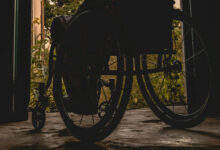 Fotoğrafta, tekerlekli sandalye kullanan engelli birey karanlık bir odadan kapıyı açarak yeşil ve gün ışığını olduğu bir bahçeye doğru çıkıyor.