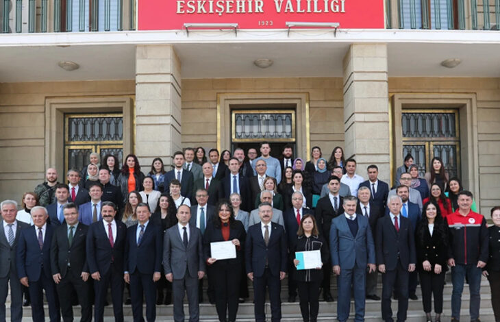 Eskişehir Valiliği tarafından düzenlenen “Ellerin Sesim Olsun” projesi kapsamında işaret dili kursunu tamamlayan memurlara belge teslim töreni Valilik önünde düzenleniyor. İşaret dili sertifikasını alan kişiler toplu fotoğraf çekimi için arkadan öne doğru ve yan yana olacak şekilde ayakta duruyorlar.