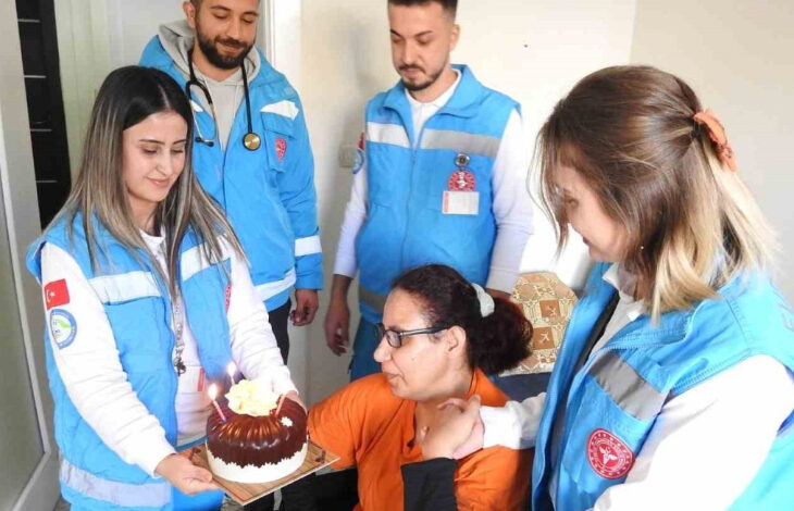 Fotoğrafta, çikolatalı bir kaplaması olan pastanın üzerinde mumlar var. Doğum günü pastası Tuba Salan’ın önüne kadar Evde Sağlık Hizmetleri ekibi tarafından getiriliyor. Tuğba'nın etrafında 4 farklı sağlık personeli var. Sağlık personellerinden ikisi kadın ikisi erkek.