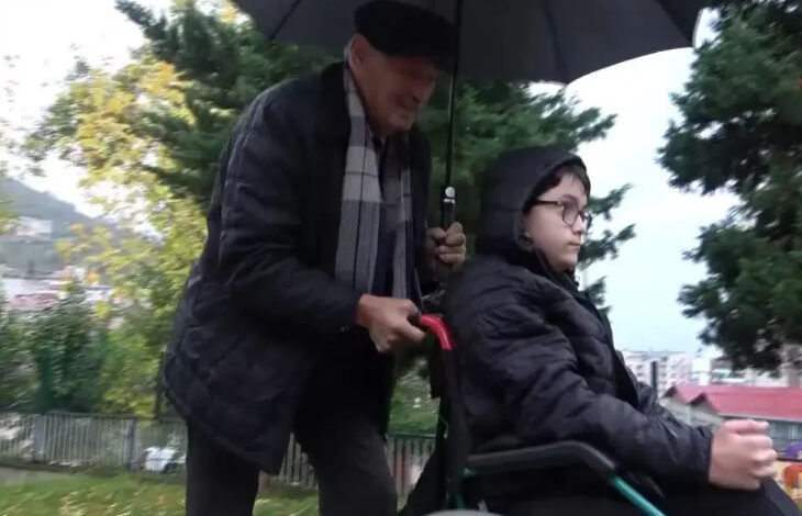 Fotoğrafta, tekerlekli sandalye üzerindeki torununu okula götüren emekli öğretmen Mustafa Sarısaray bir eliyle şemsiye tutuyor, diğer eliyle ise tekerlekli sandalyeyi sürüyor.