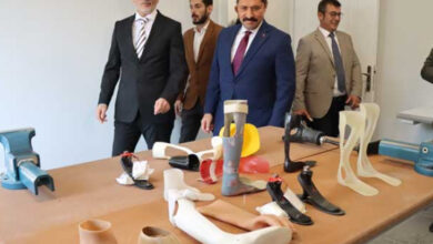 Fotoğrafta, masa üzerinde yer alan farklı boyutta, farklı renklerde olan protez ürünleri takım elbise giymiş kalabalık bir erkeklerden oluşan grup inceliyor.