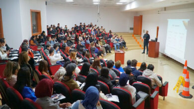 Fotoğrafta, Mersin Üniversitesi İletişim Fakültesi öğrencileri engelli farkındalığı üzerine yapılan semineri izliyor. Sahnede anlatımı gerçekleştiren bir akademisyen var.