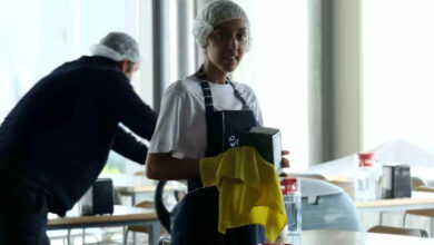 Masaların ve sandalyelerin olduğu bir odada, masaların üzerinde peçete kutuları, su şişeleri yer alıyor. Fotoğraf makinesine doğru bakın genç bir kadın elindeki peçete kutusunun tozunu bez ile temizliyor.