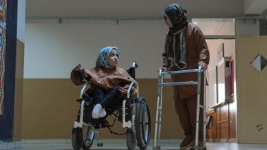 Serebral palsili öğretmeni Senem Arıcı, önündeki yürütece tutunarak ayakta duruyor ve yanında akülü tekerlekli sandalye kullanan engelli öğrencisi var.