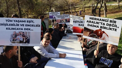 Fotoğrafta, etkinlik kapsamında bir araya gelen engelli bireyler uzun bir masanın etrafında otururken, ellerinde çeşitli pankartları tutuyorlar.