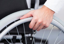 Tekerlekli sandalye üzerinde oturan engelli bireyin, sandalyeyi hareket ettirmek için teker bölümünde yer alan itme demirlerini tuttuğu görülmektedir.