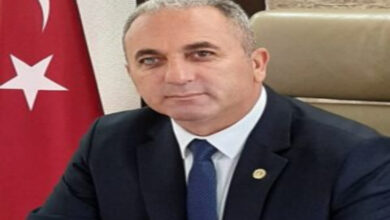 Fotoğrafta, Adem Kuyumcu gri bir koltuk üzerinde oturuyor ve arkasında ise Türk Bayrağı var.