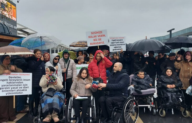 Fotoğrafta, engelli bireyler yağmurlu bir havada tekerlekli sandalyeleri üzerinde oturarak ve bazılarının elinde pankartlarla birlikte basın açıklamasına katılıyorlar.