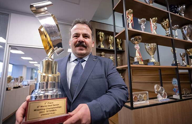TİESF’in yeni başkanı Dursun Gözel madalyaların ve kupaların olduğu bir alanda büyük bir kupayı elinde tutuyor.