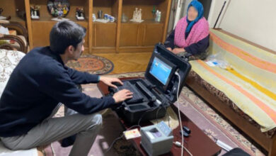 Fotoğrafta, koltukta oturan yaşlı kadın karşı tarafında oturan ve masanın üzerine bilgisayar benzeri bir cihaz katan görevli tarafından kimliği yenileniyor.