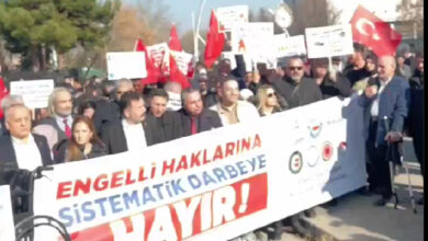 “Engelli haklarına sistematik darbeye hayır” yazılı bir pankartın arkasında engelli bireyler ve sivil toplum kuruluşu temsilcileri bayrak ve pankartlar tutarak basın açıklamasına destek veriyor.