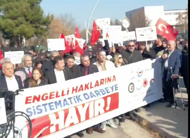 “Engelli haklarına sistematik darbeye hayır” yazılı bir pankartın arkasında engelli bireyler ve sivil toplum kuruluşu temsilcileri bayrak ve pankartlar tutarak basın açıklamasına destek veriyor.