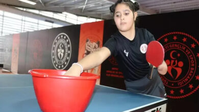 Fotoğrafta, genç bir kız masa tenisi oynarken görülüyor. Siyah bir spor tişörtü giymiş ve elinde kırmızı bir raket tutuyor. Masa tenisinin oynandığı ortamda, masanın üzerinde kırmızı bir plastik kova bulunuyor ve kız elini içine uzatıyor. Arka planda Gençlik ve Spor Bakanlığı logosu ve çeşitli spor temalı görsellerin yer aldığı bir duvar var. Spor salonunun tavanı metal konstrüksiyonlarla kaplı ve geniş bir alan izlenimi veriyor. Fotoğraf, genç sporcunun masa tenisi antrenmanı yaptığı bir anı yansıtıyor.