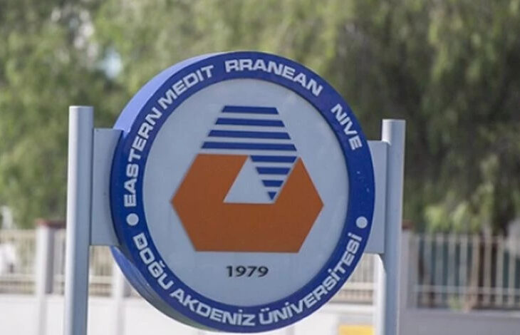 Bu görselde, Doğu Akdeniz Üniversitesi'ne (DAÜ) ait bir tabela veya logo yer almaktadır. Logo, yuvarlak bir çerçeve içerisinde tasarlanmış ve üst kısmında İngilizce "Eastern Mediterranean University" (tam olarak "Eastern Mediterranean University" ibaresinin tamamlanmamış olduğu anlaşılıyor) yazısı yer alıyor. Alt kısmında ise Türkçe "Doğu Akdeniz Üniversitesi" ibaresi bulunuyor. Logonun merkezinde, turuncu bir altıgen şekil ile mavi renkli, şeritler şeklinde bir grafik tasarım yer alıyor. Alt kısımda, üniversitenin kuruluş yılı olan "1979" yazısı bulunmakta. Logo, mavi, turuncu ve beyaz renk temalarını içeriyor ve arka planda bulanık bir açık hava sahnesi görülüyor.