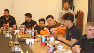 Fotoğrafta uzun bir yemek masası etrafında oturan bir grup erkek görülüyor. Masanın üzerinde yemek tabakları, içecek kutuları ve şişeleri, ekmek sepeti gibi çeşitli yiyecek ve içecekler yer almakta. Katılımcılardan bazıları birbirleriyle sohbet ediyor ya da yemekle meşgul. Arka planda ise ayakta duran bir kişi dikkat çekiyor. Masadaki kişiler genellikle spor kıyafetleri giymiş, üzerlerinde logolar bulunuyor. Ortam samimi ve bir toplantı ya da yemek etkinliği havası veriyor.