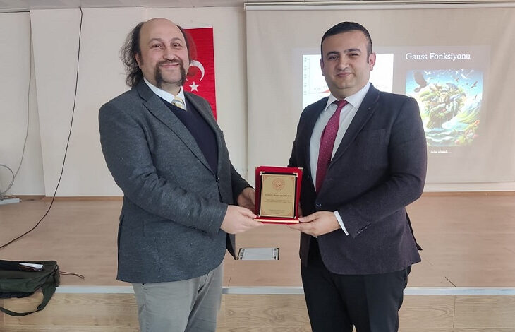 Fotoğrafta, iki erkek birey bir sahnede yan yana duruyor ve ellerinde bir plaket tutuyorlar. Soldaki kişi, ceket ve kravatla resmi bir kıyafet giymiş, gülümseyerek objektife bakıyor. Sağdaki kişi de takım elbise ve kırmızı kravat giymiş, aynı şekilde gülümseyerek plaketin bir tarafını tutuyor. Arkada bir Türk bayrağı ve projeksiyon perdesi üzerinde "Gauss Fonksiyonu" yazısı ve görsel yer alıyor. Mekan, bir toplantı veya konferans salonunu andırıyor.