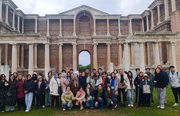 Fotoğrafta, büyük bir grup insan tarihi bir yapının önünde poz veriyor. Arka planda, sütunları ve kemerli girişleriyle dikkat çeken, antik bir mimariye sahip büyük bir yapı yer alıyor. İnsanlar kışlık kıyafetler giymiş, bazıları atkı ve bereler takmış. Grupta farklı yaş gruplarından bireyler bulunuyor; bazıları gülümseyerek poz verirken, bazıları daha ciddi bir ifadeye sahip. Görme engelli bireyler beyaz baston kullanıyor. Fotoğraf, tarihi ve kültürel bir gezi etkinliği sırasında çekilmiş gibi görünüyor.