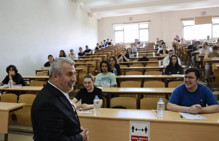 Fotoğrafta, bir üniversitenin sınıfında öğrenciler ve öğrencilerin önündeki boş alanda ders anlatan bir üniversite hocası var.