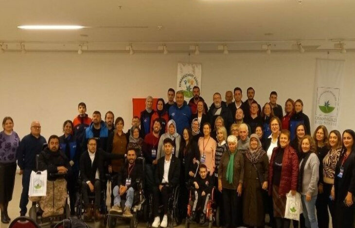 Fotoğrafta, bir grup insan geniş bir salonda bir araya gelmiş şekilde görülüyor. Çeşitli yaş gruplarından bireyler, hem engelli bireyler hem de onların destekçileri olarak, birlikte poz vermişler. Grupta tekerlekli sandalyedeki kişiler de dikkat çekiyor ve toplumun her kesiminden bireylerin bir araya geldiği bir etkinlik ortamı olduğu anlaşılıyor. Arka planda Osmangazi Kent Konseyi ve Engelliler Meclisi'nin logoları yer alırken, Türk bayrağı da karede görünmekte. Etkinliğin amacı, "Afetlerde Engelsiz Birlik" gibi bir başlıkla engellilerin afet durumlarında yaşadığı zorluklara yönelik farkındalık yaratmak ve çözüm üretmek olarak öne çıkıyor. Toplumsal dayanışma ve birlik mesajı veren anlamlı bir anı yansıtıyor.